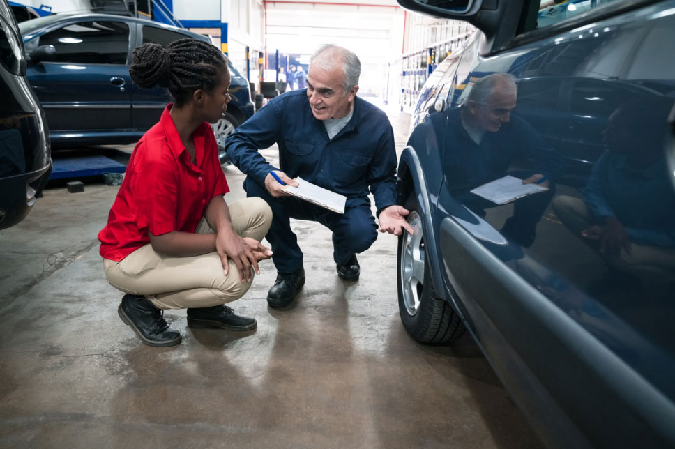 A woman and a mechanic squat down next to her car in a garage.